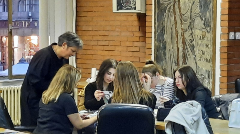 Photo of a meeting with researchers at University of Belgade, students are sat around a table talking to Lynda who is standing up